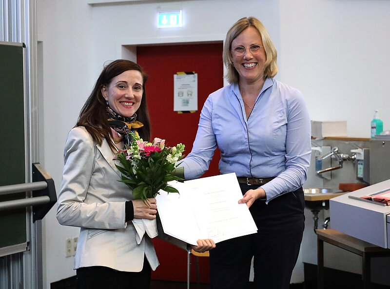 Two women stand smiling at the camera. The woman on the left holds a bouquet of flowers she has just received, and both women hold a certificate between them. 