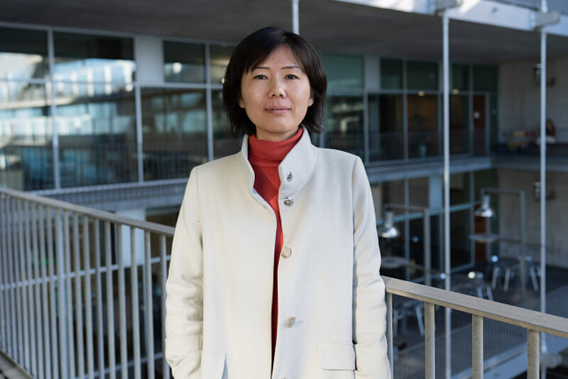 An image of a professor standing in the centre of a photograph. She is leaning against a railing overlooking a courtyard. 