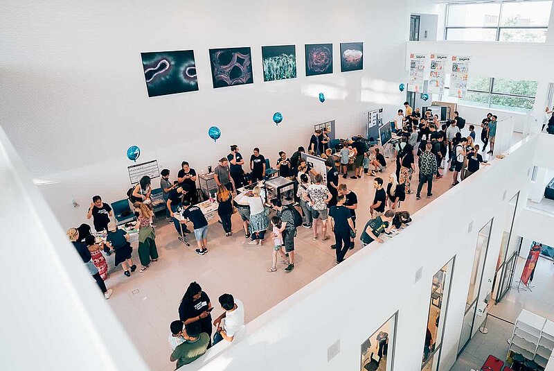 Large crowds of people cluster around a series of tables, with blue balloons floating in the air above them. To the right, 3 banners with comics on them hang from the walls. Many people are interacting with activities or items on the tables or watching demonstrations by scientists. 