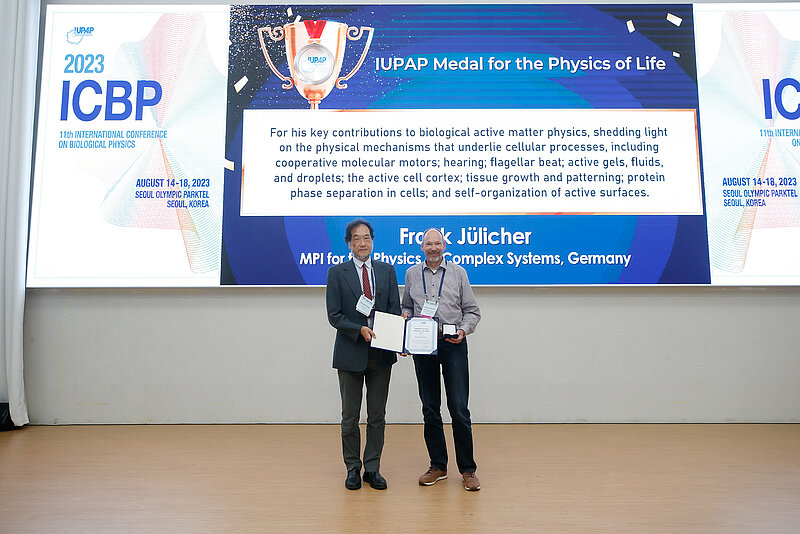 Two people on the foreground both holding a certificate of the pirze and on the background a slide with explanation of his contribution for the prize