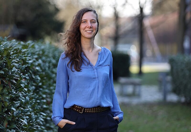 A woman in a blue shirt is smiling at the camera, to the left of her are some bushes and to the right we can see a lawn