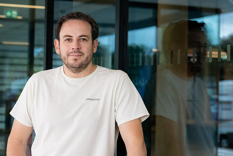 A man in a white t-shirt stands before a glass window smiling 