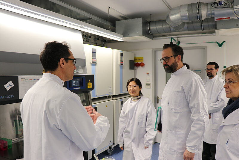 People standing in the laboratory wearing white labcoats. A man is in the front explaning to the rest of people.
