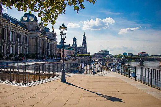 View from standing on the Brühlsche Terrasse in Dresden towards the Academy of Fine Arts, the Dresden Cathedral, and the Semper Opera (on the left hand side) and the river Elbe (on the right hand side). In the foreground a treetop hanging into the picture and a gas lantern.