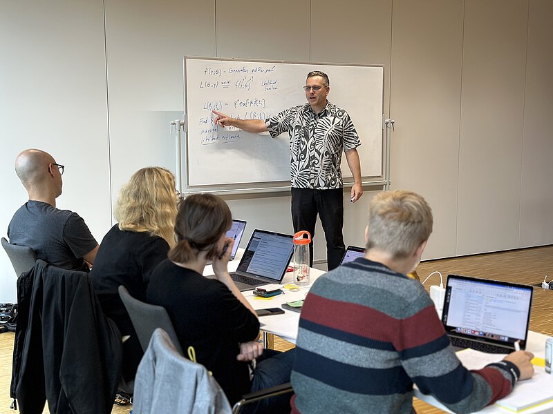 Prof. Justin Bois is at the background pointing to a white board, in which some equations about statistics, at the foreground attendants of the workshop are shown