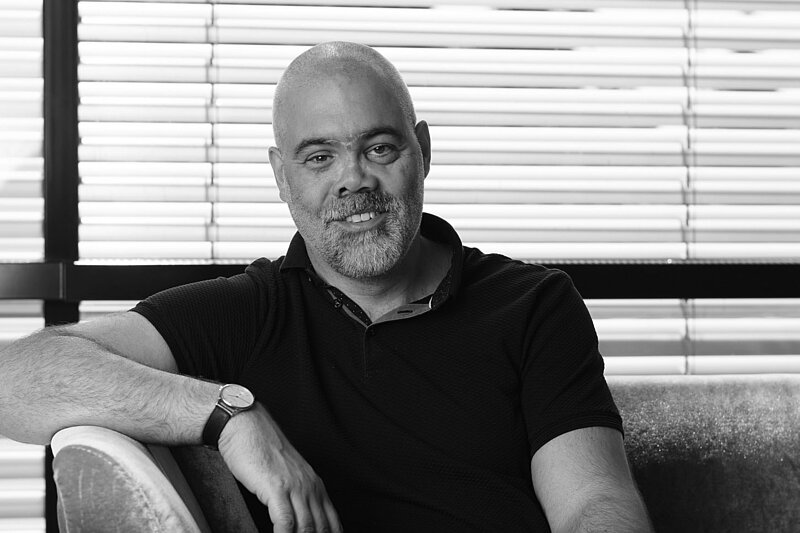 A black and white image depicts Professor Jochen Guck sitting on a couch smiling at the camera. His right arm is leaning on the armrest of the couch and he wears a black t-shirt. 