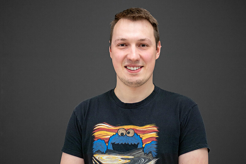 A man stands smiling in the centre of the image wearing a black t-shirt with a colourful pattern on it