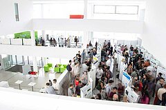 A scientific poster session in the open atrium of white scientific institute, with a large crowd of people, seen from above