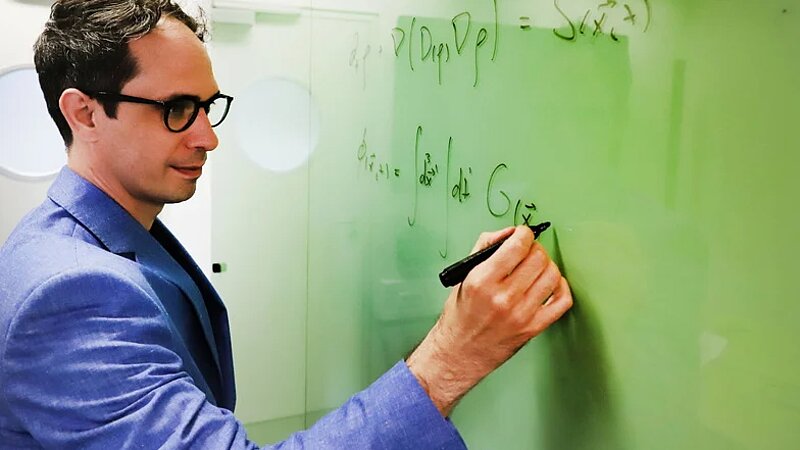 A man with glasses stands on the left hand side of photograph writing a series of equations on a whiteboard. 