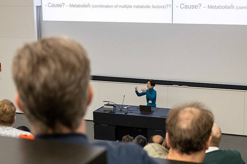 A professor stands at a table with her arms outstretched, explaining a concept to an audience in the foreground, we the back of the audience's heads.  