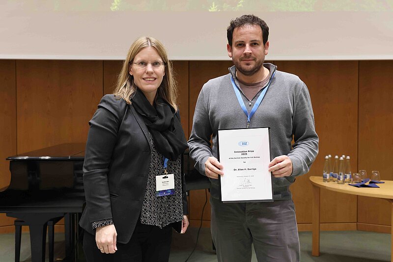 A woman stands on the left wearing glasses and a black outfit offering a certificate to the main on the right wearing grey. Both are smiling at the camera. 