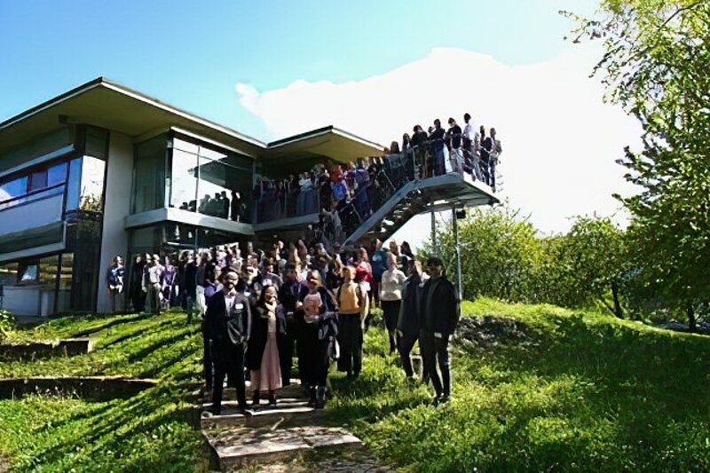 A group of people stand on a green hill with the sun and large glass building behind them. 