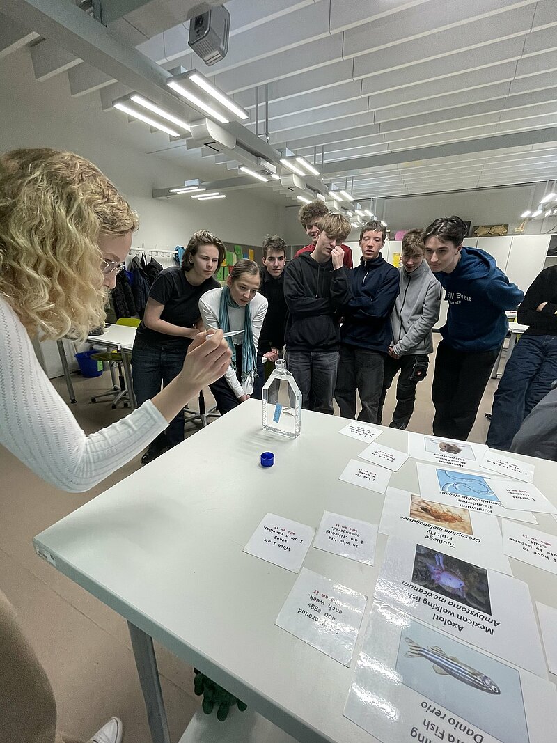 A group of students stand around a table, while a PhD researcher teaches them. She holds a plastic pipette in her hand. 