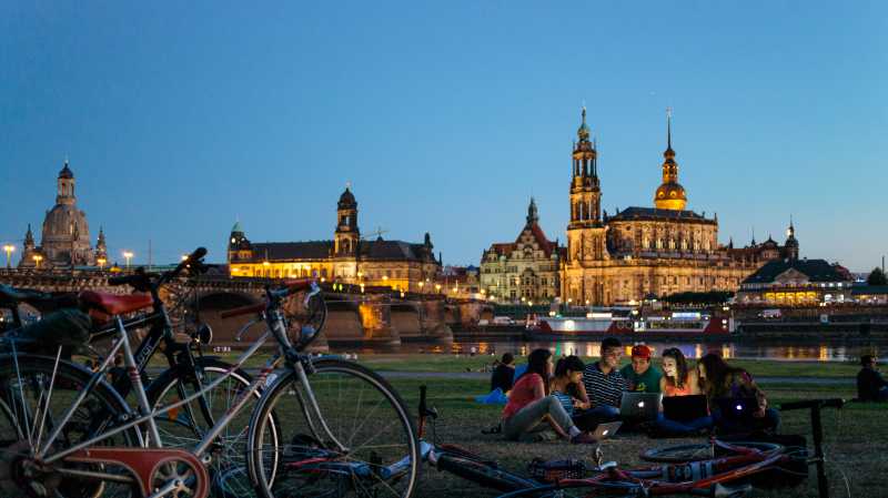 Young people sitting in the grass by the river Elbe with the historic city center of Dresden in the background.