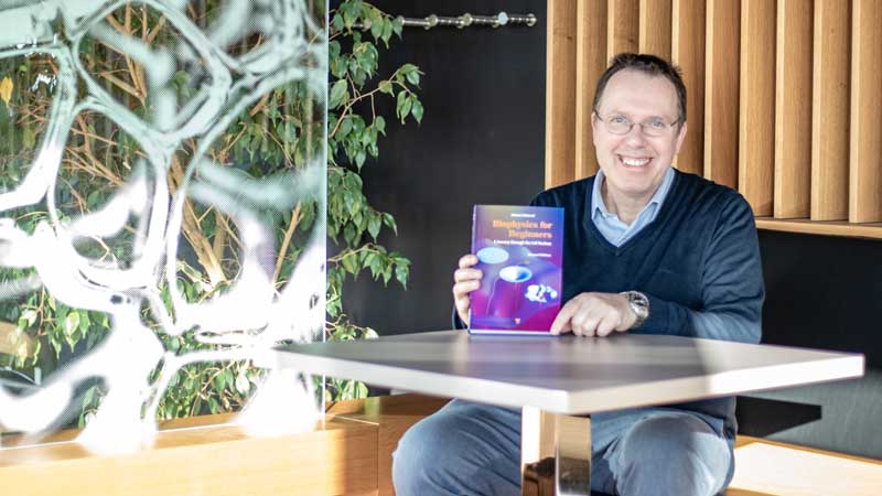 The image shows Prof. Helmut Schießel sitting at a table with the 2nd edition of his textbook "Biophysics for Beginners: A Journey Through the Cell Nucleus" © Hagen Gebauer (TUD)